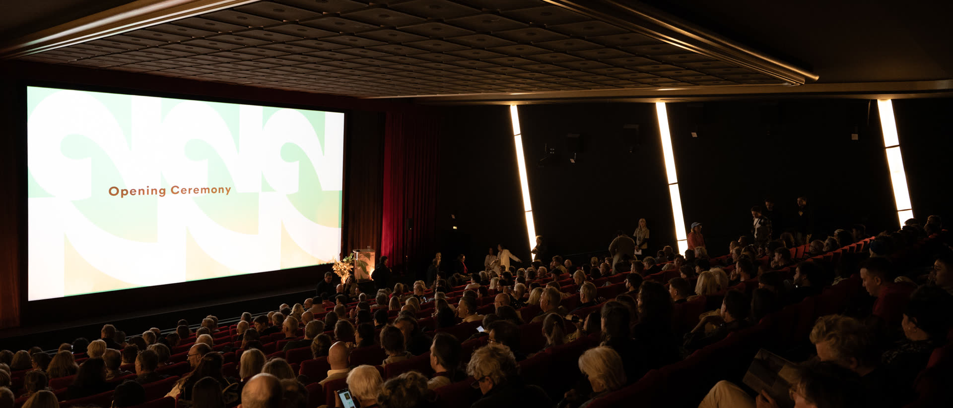 A full cinema auditorium, the audience seated and the screen lit